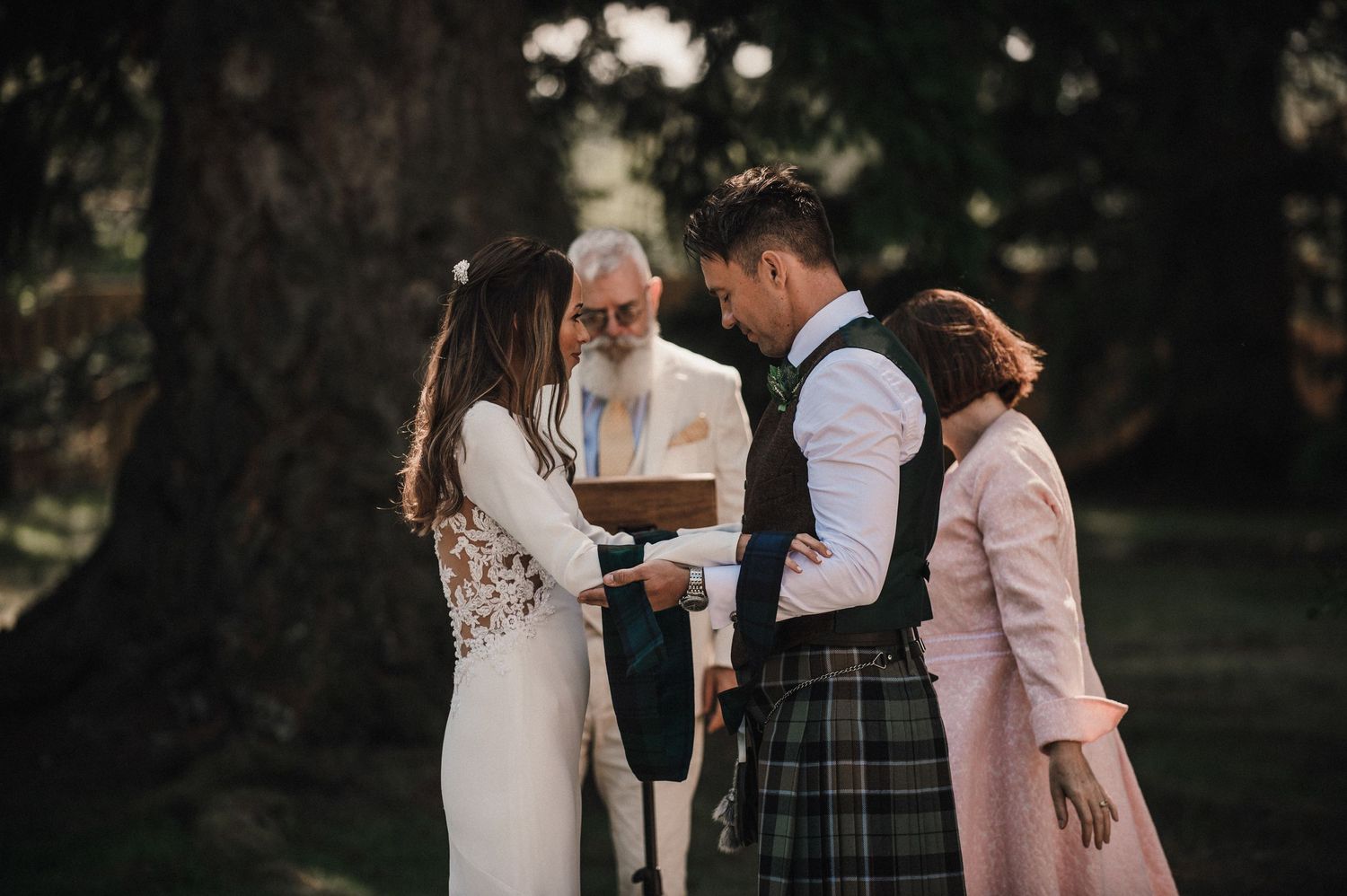 A couple participates in their wedding ceremony with an officiant under tall trees.