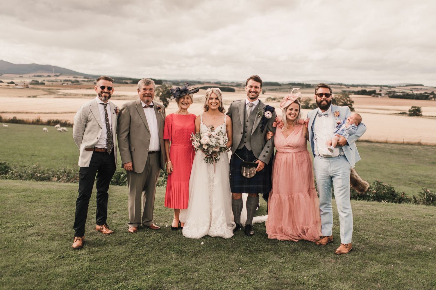 Wedding party members in bright colors and kilts pose together on grassy hilltop.