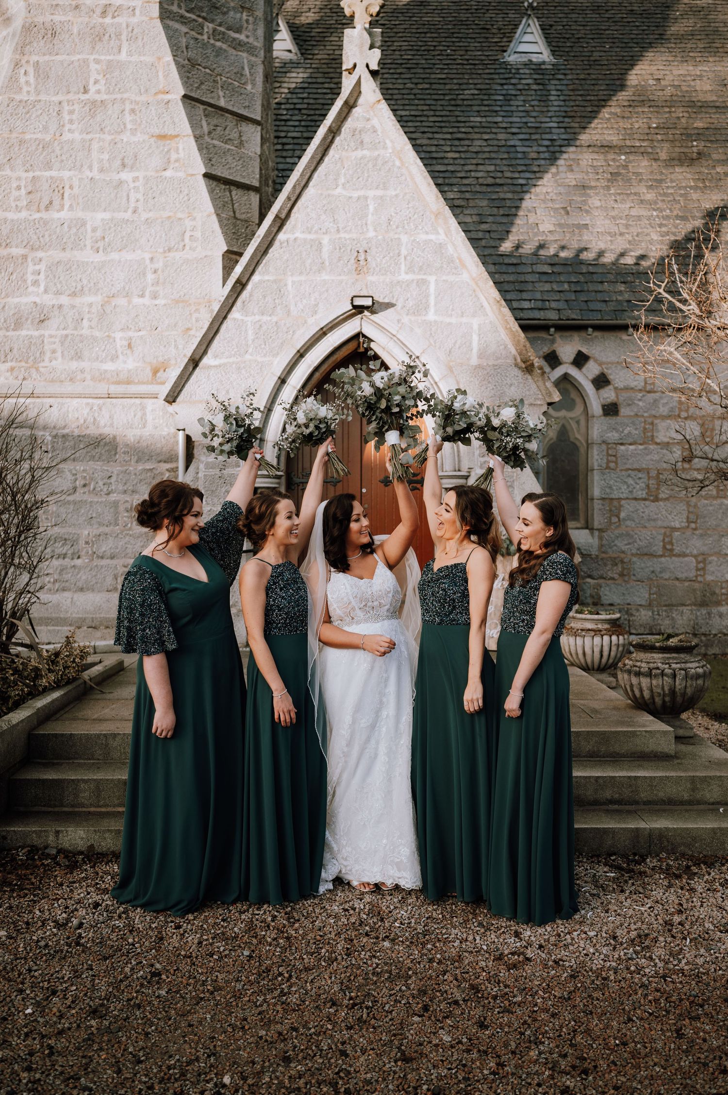 Wedding party in emerald dresses celebrate joyfully outside gothic church entrance.
