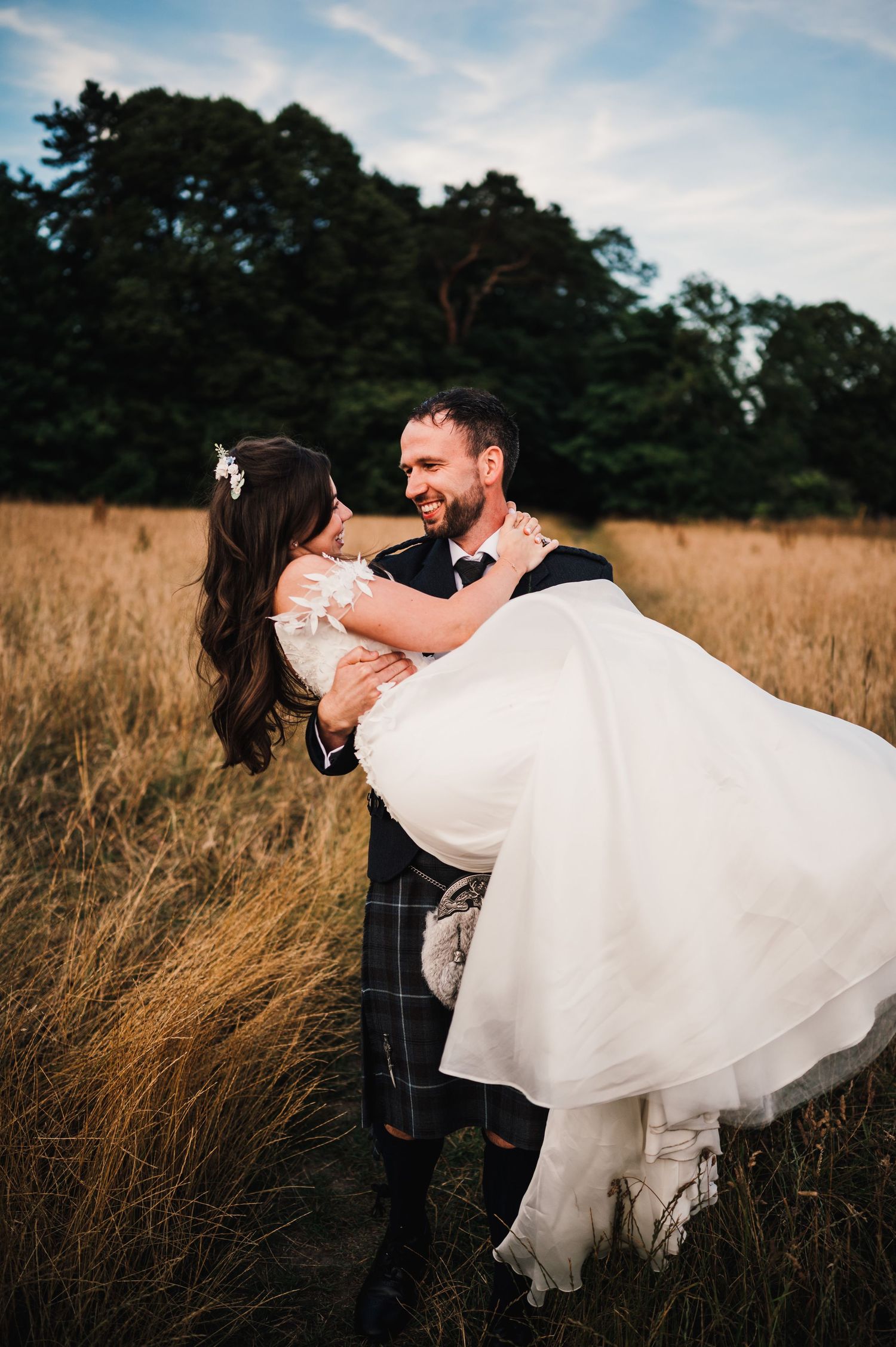 A groom in traditional Scottish attire carries his bride through a sunlit field.