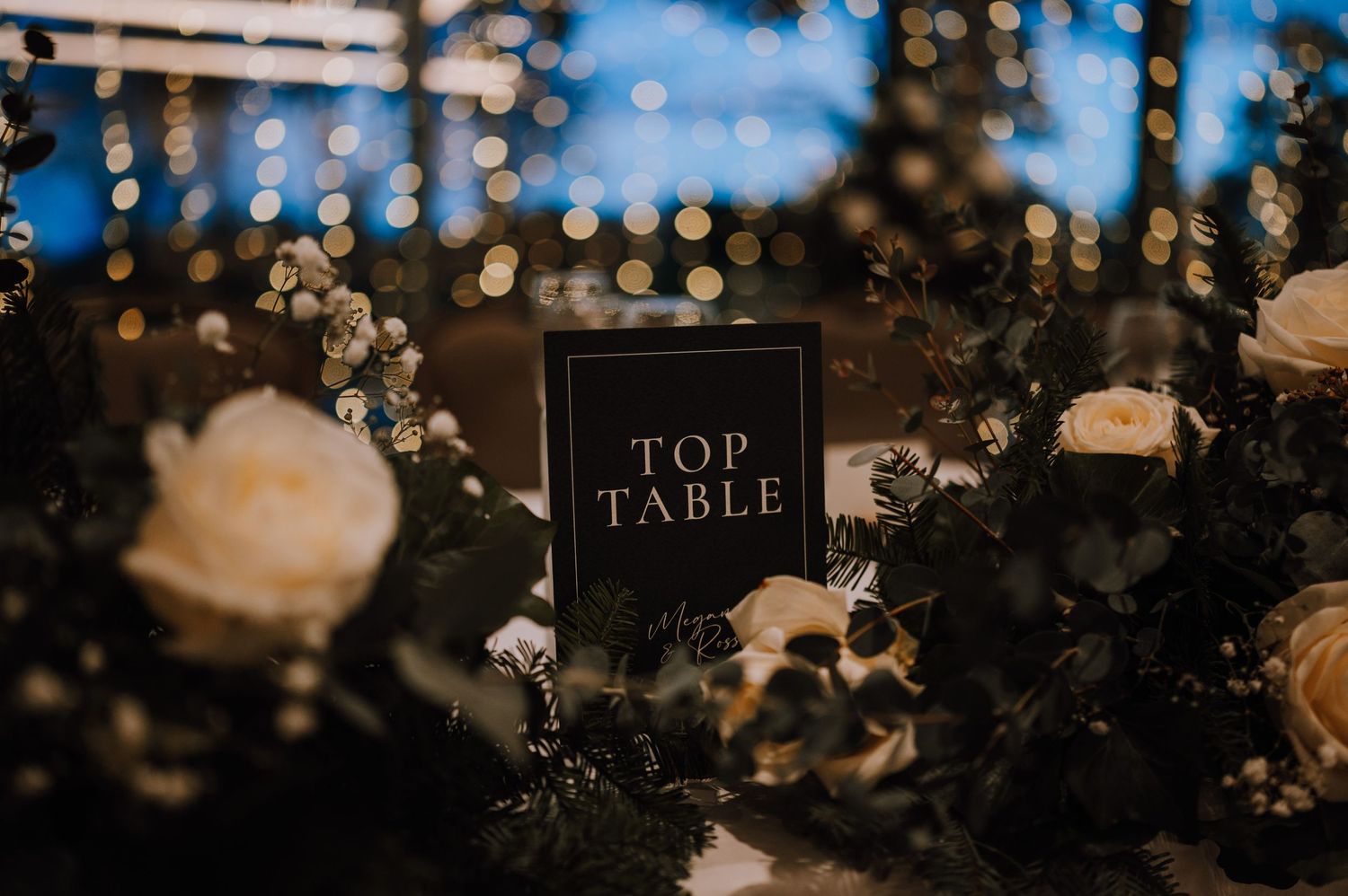 Top table sign nestled among white roses and greenery at evening reception.