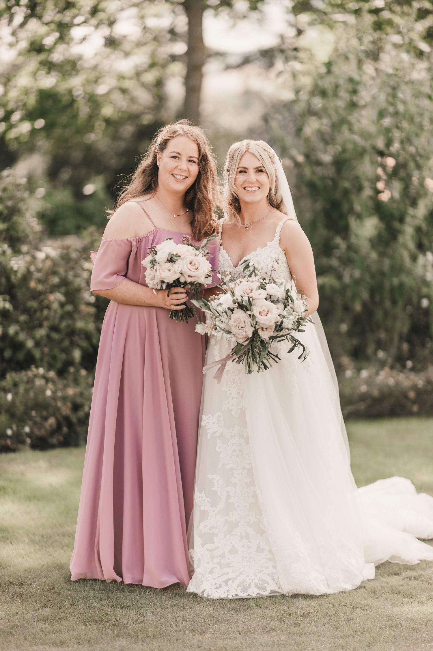 Outdoor wedding photography featuring bridesmaid in dusty rose gown and bride in lace dress with matching bouquets.