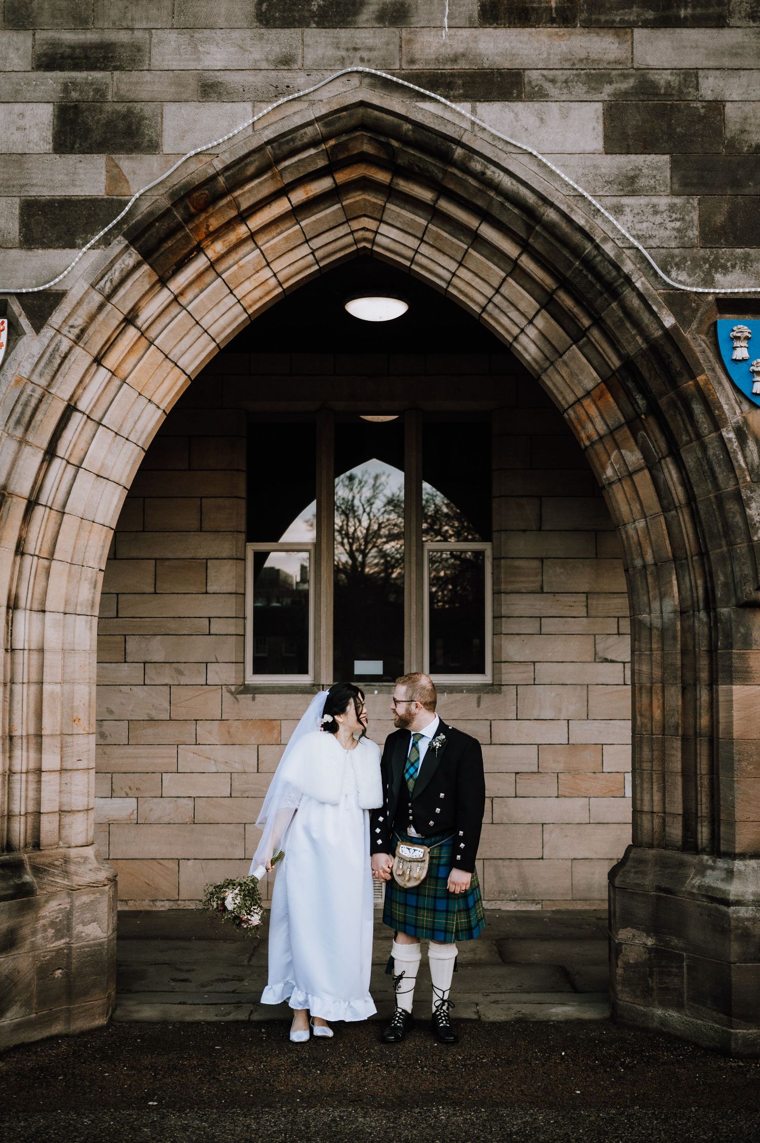 Couple poses under stone church archway in winter wedding attire with Scottish details.