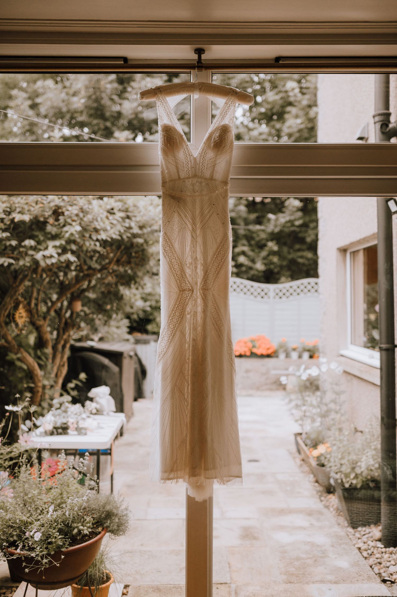 An elegant white wedding dress hangs in front of a window with garden views.