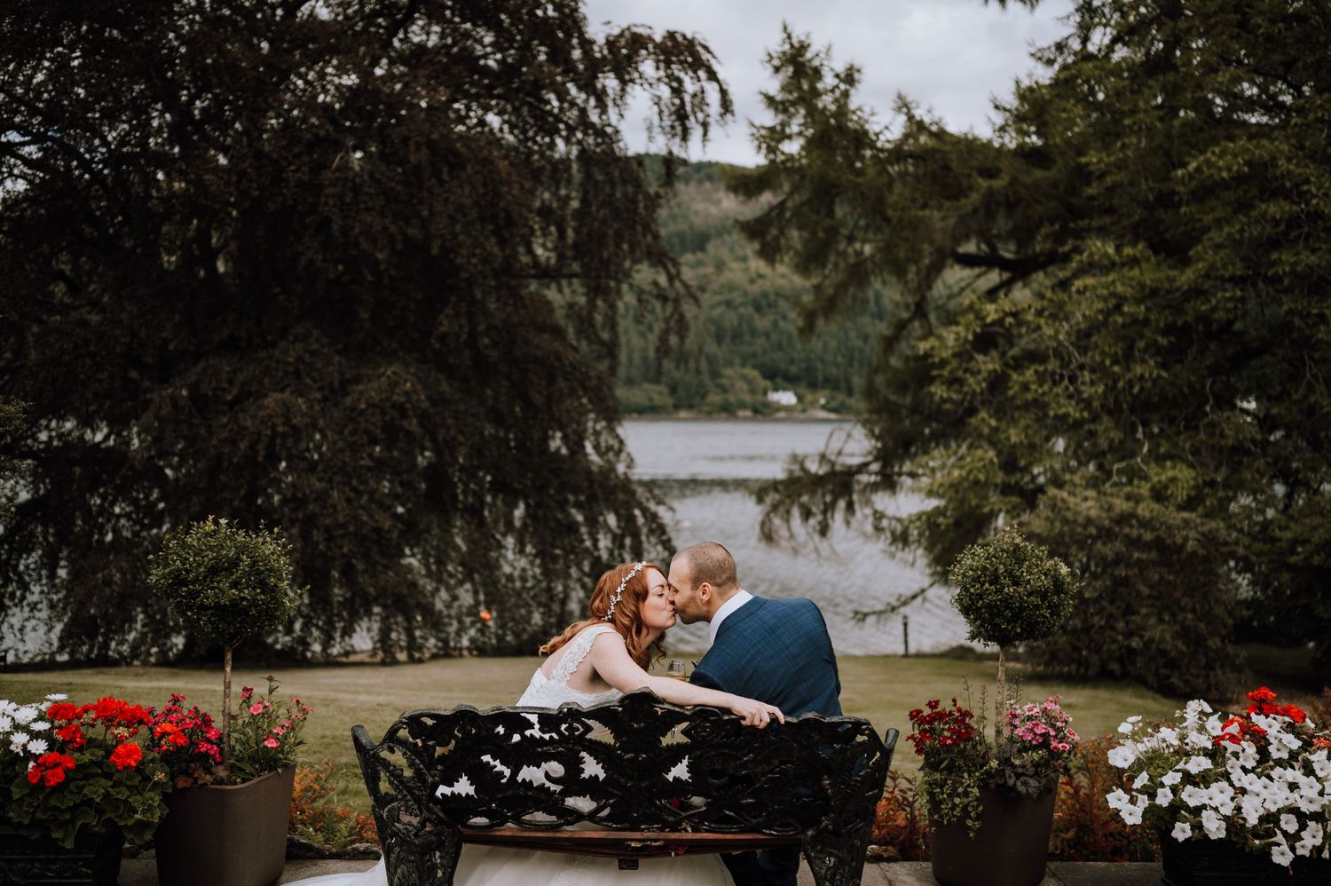 Couple shares intimate moment on garden bench surrounded by colorful flowers with lake view.