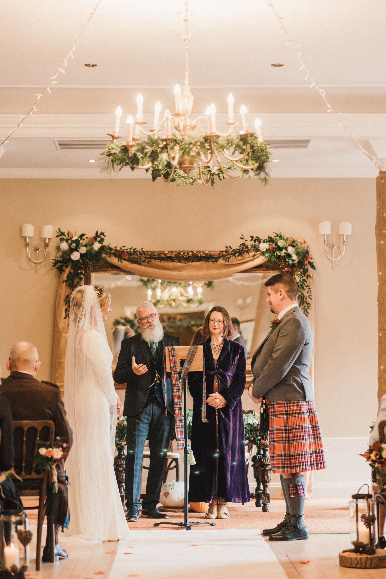 A Scottish wedding ceremony under an elegant chandelier with floral arch decorations and traditional Highland dress.