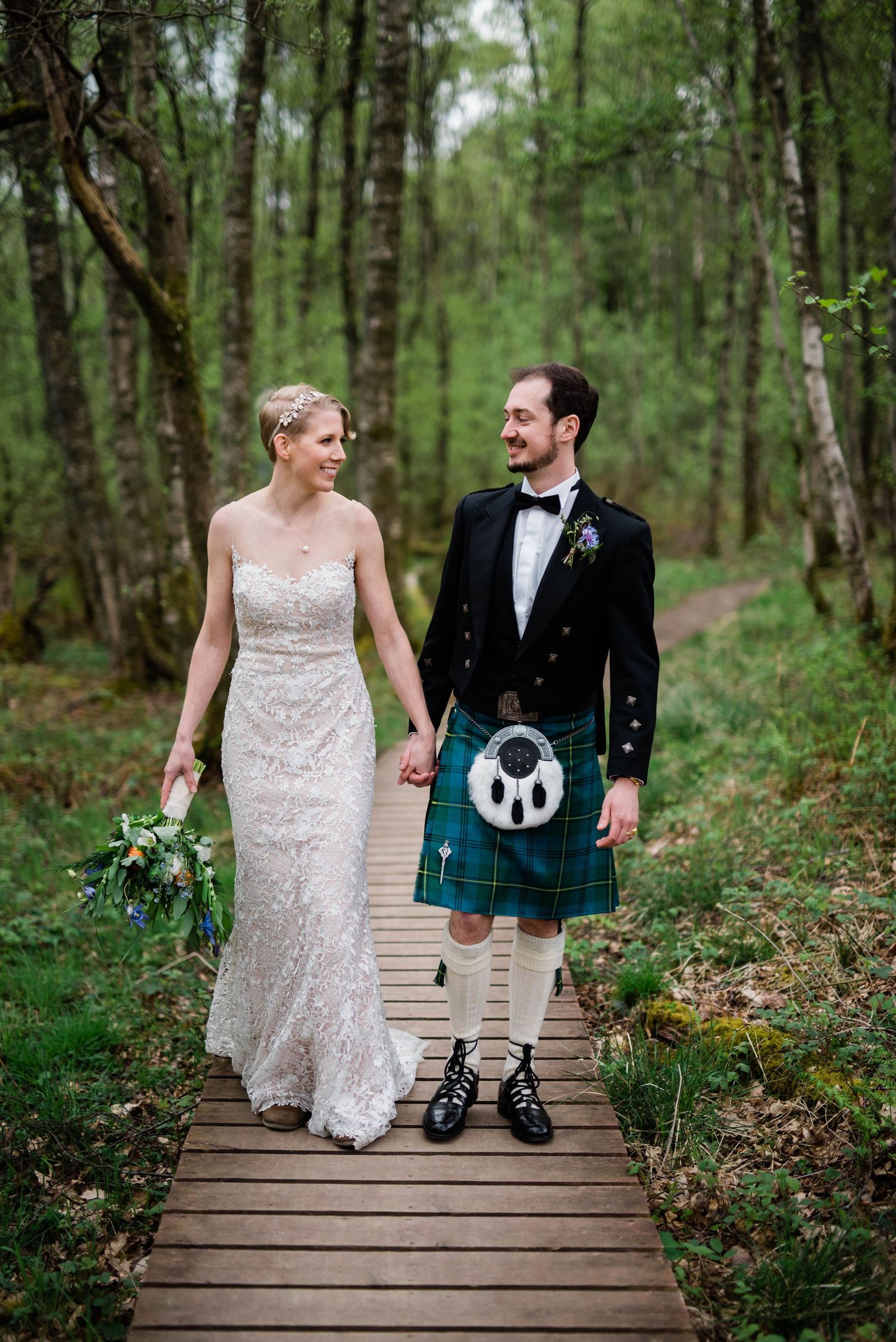 A couple walks along a wooden boardwalk through a forest, with the groom wearing a traditional Scottish kilt.