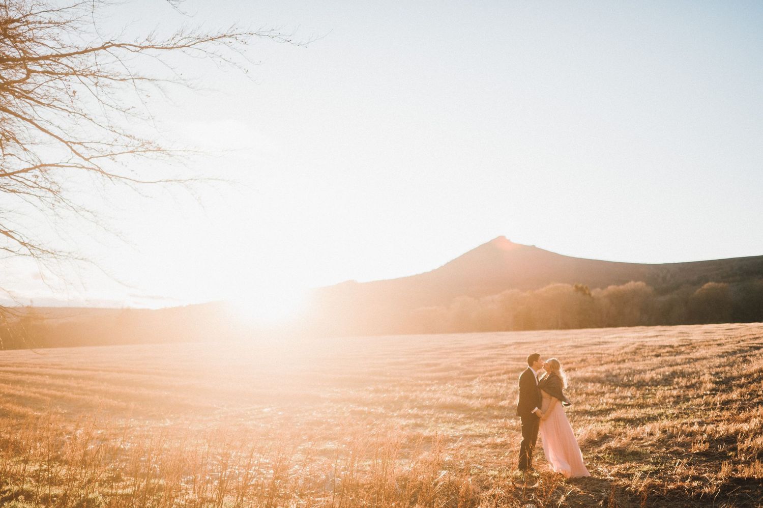 A romantic couple stands in a golden field at sunset with mountains in the background.