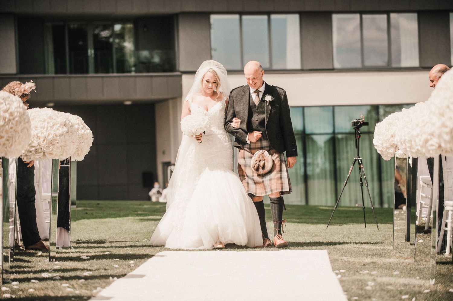 A wedding couple walks down the aisle with the bride in white and groom wearing a traditional Scottish kilt.
