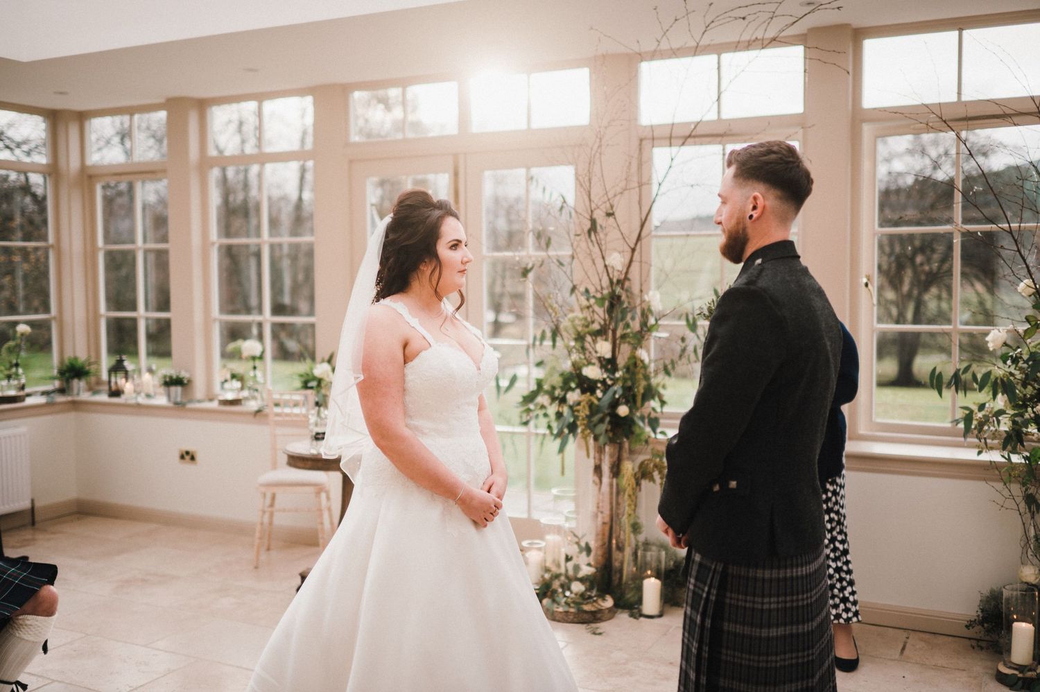 A Scottish wedding couple shares a moment in a bright conservatory with large windows.