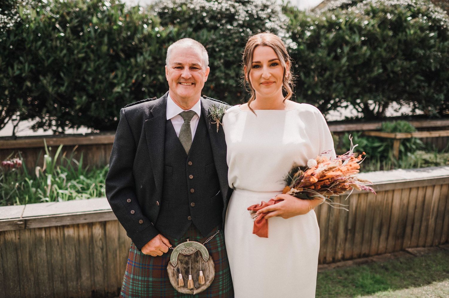 A Scottish wedding couple poses outdoors, with the bride holding an autumn-colored bouquet.