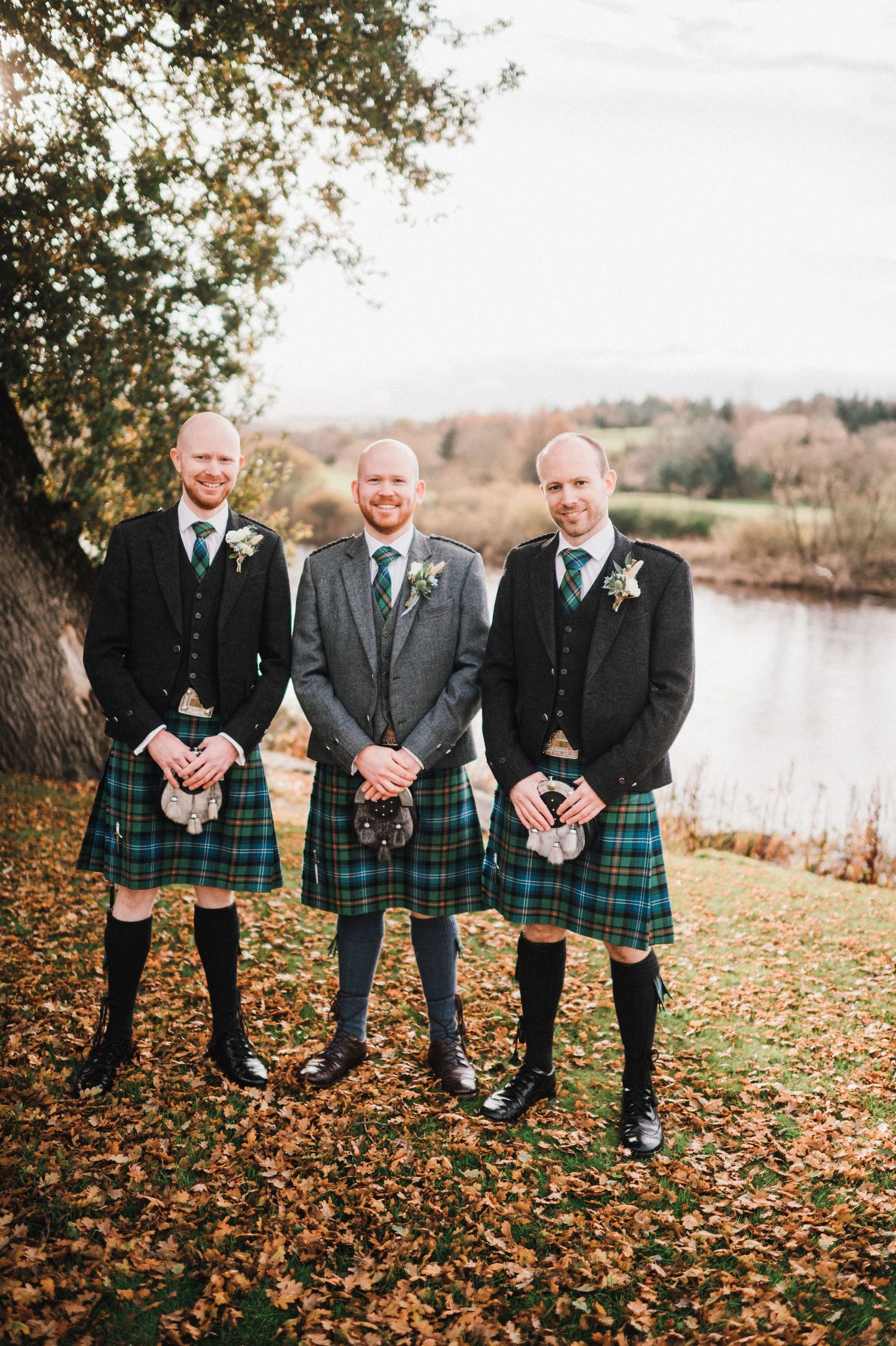 Three men in matching kilts and formal wear pose beneath a tree near a scenic lake.