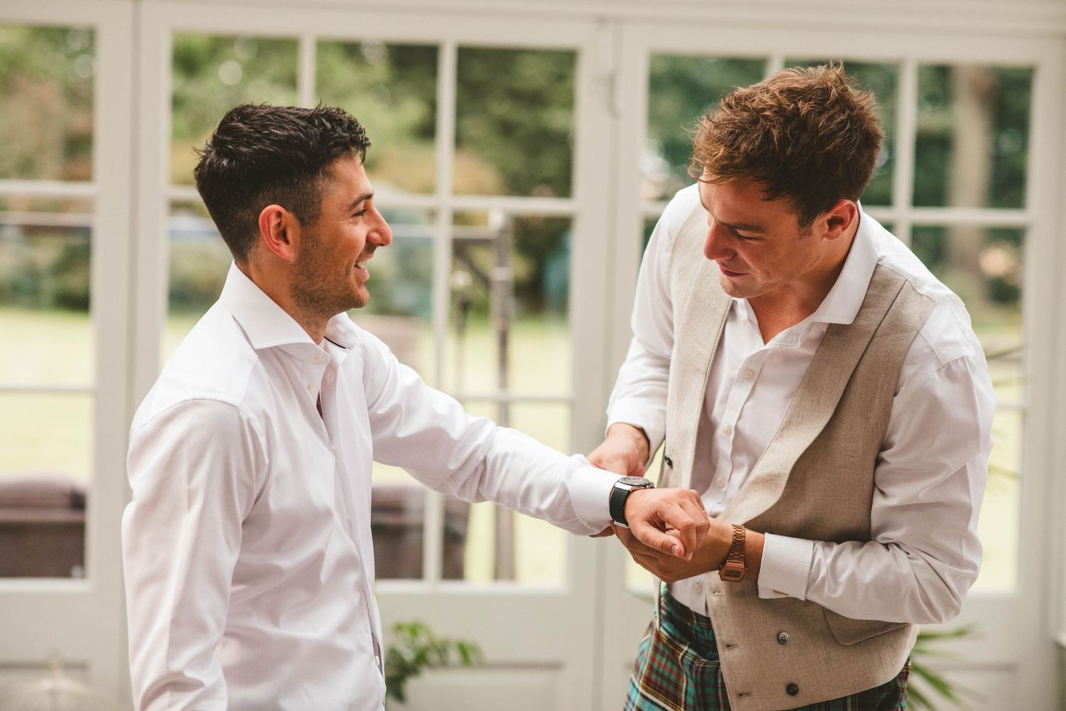 Two men in formal attire examining something in hands by sunlit windows.