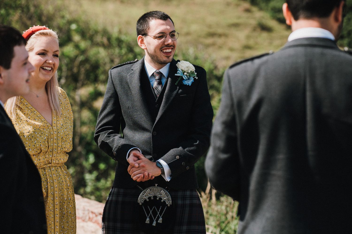 A wedding ceremony takes place outdoors with guests and groomsmen in traditional Scottish attire.