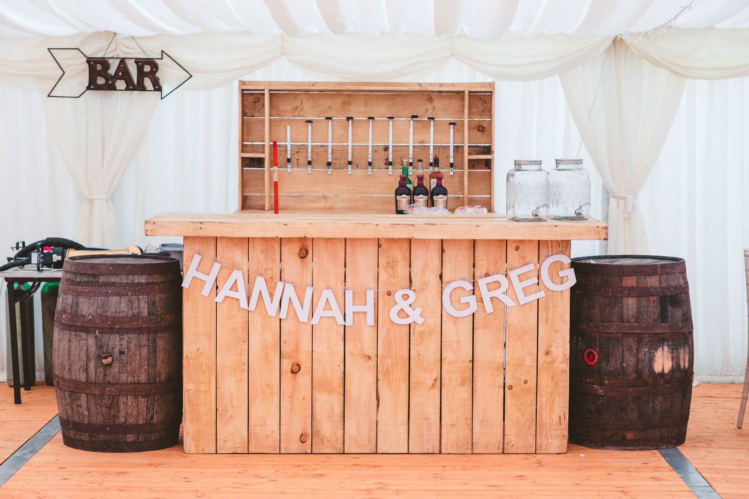 A rustic wooden wedding bar setup with whiskey barrels and string lights in a white marquee tent.
