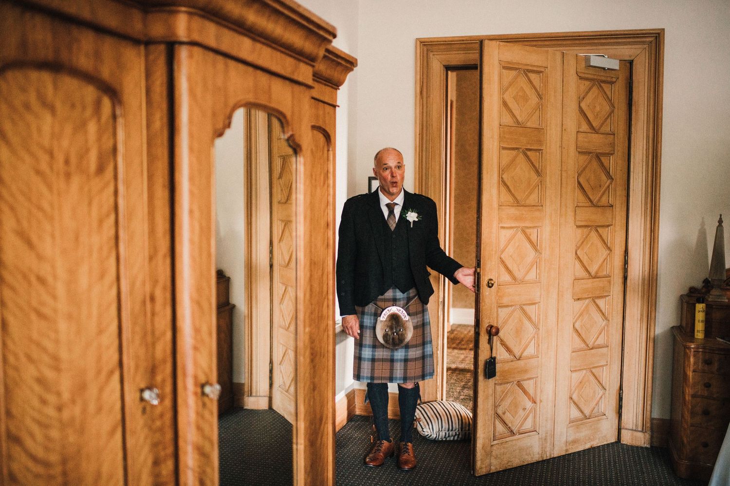 A person in traditional Scottish kilt attire stands in a room with ornate wooden furniture.