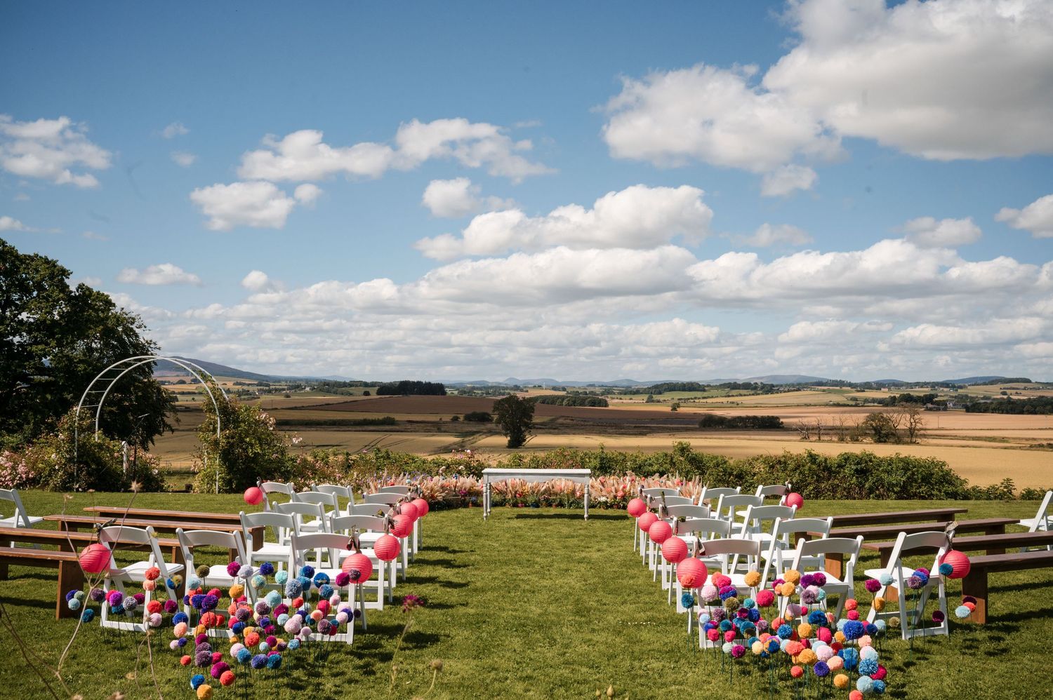 Panoramic view of an outdoor wedding ceremony space with white chairs and pink accents on a grassy hillside.