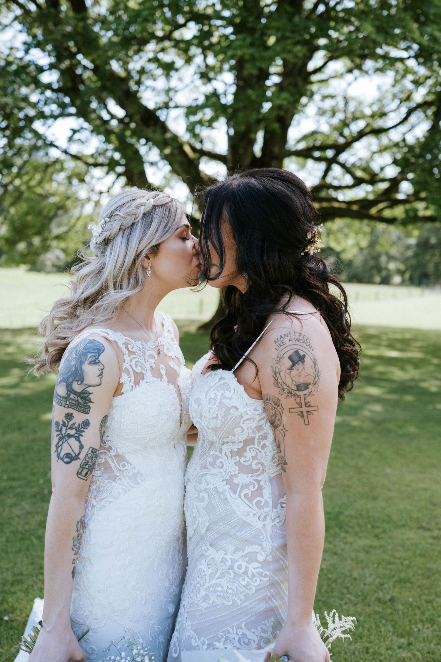 Two brides in white lace wedding dresses sharing an intimate moment, both with visible tattoos.