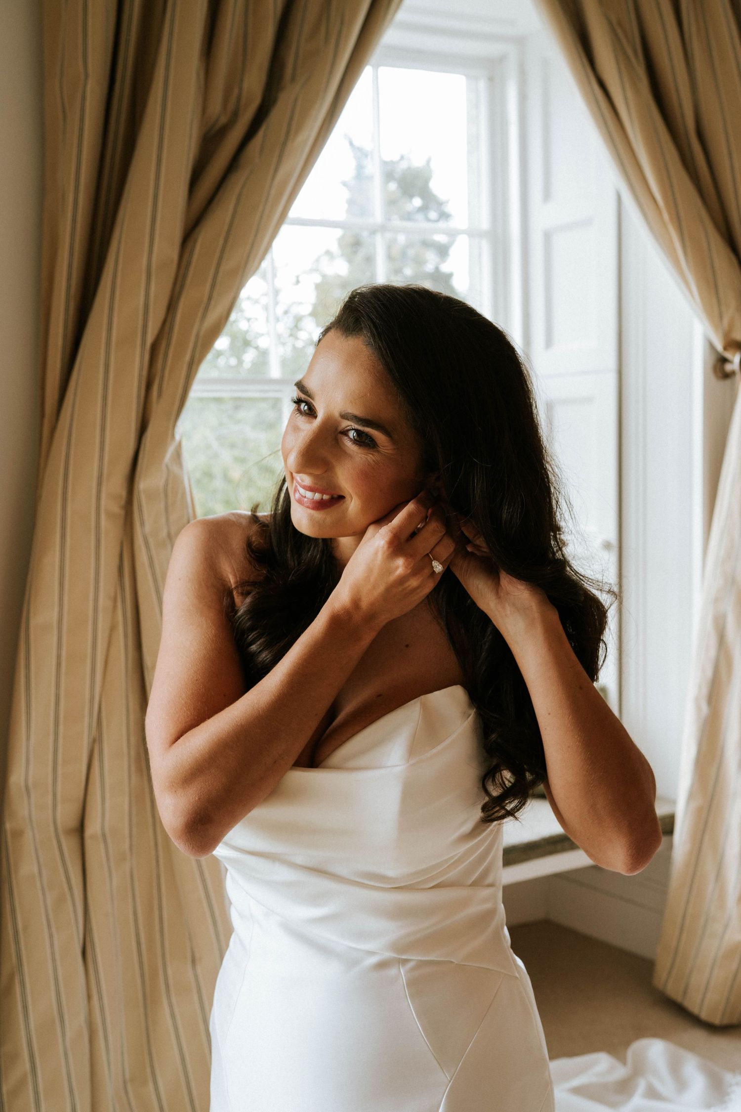 A person in a white dress adjusts their earrings near a window with striped curtains.