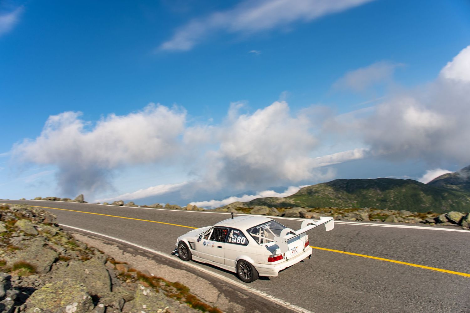 White race car with spoiler parked on a mountain highway under a dramatic blue sky with clouds.