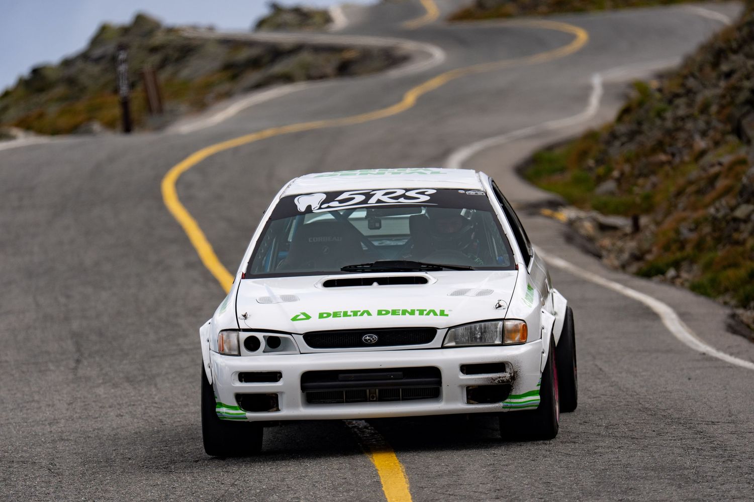 White Subaru rally car with Delta Dental sponsorship racing on a winding mountain road at Pikes Peak hillclimb.