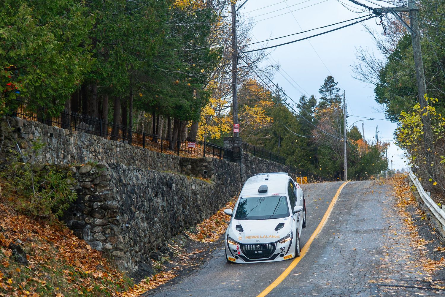 White car navigating steep mountain road with rock wall and autumn foliage.