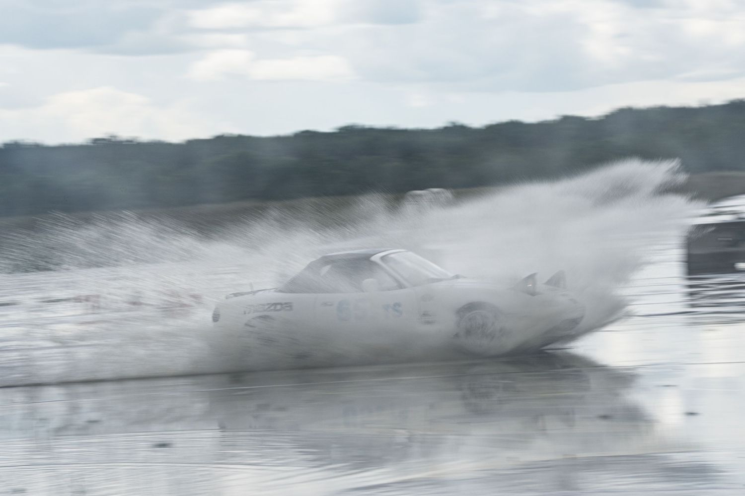 A race car speeds through a massive water splash on a wet track, creating dramatic spray under cloudy skies.