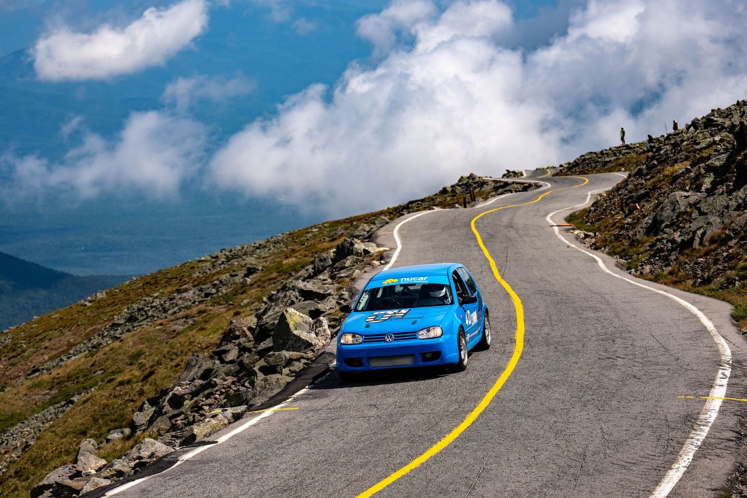 Blue rally car racing up a winding mountain road under dramatic cloudy skies on a sunny day.