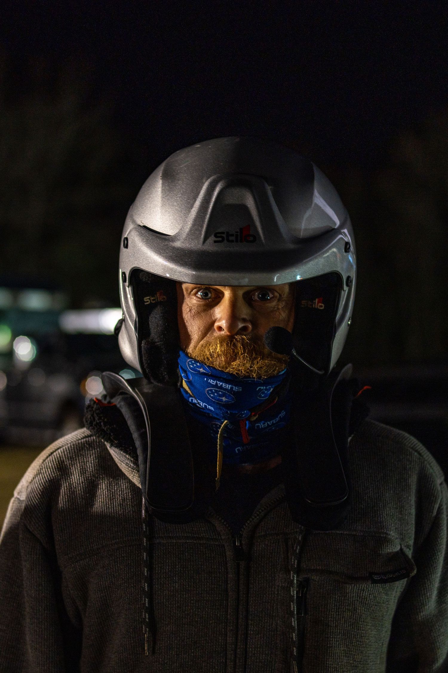 Rally driver wearing protective helmet and neck gaiter in low light conditions.