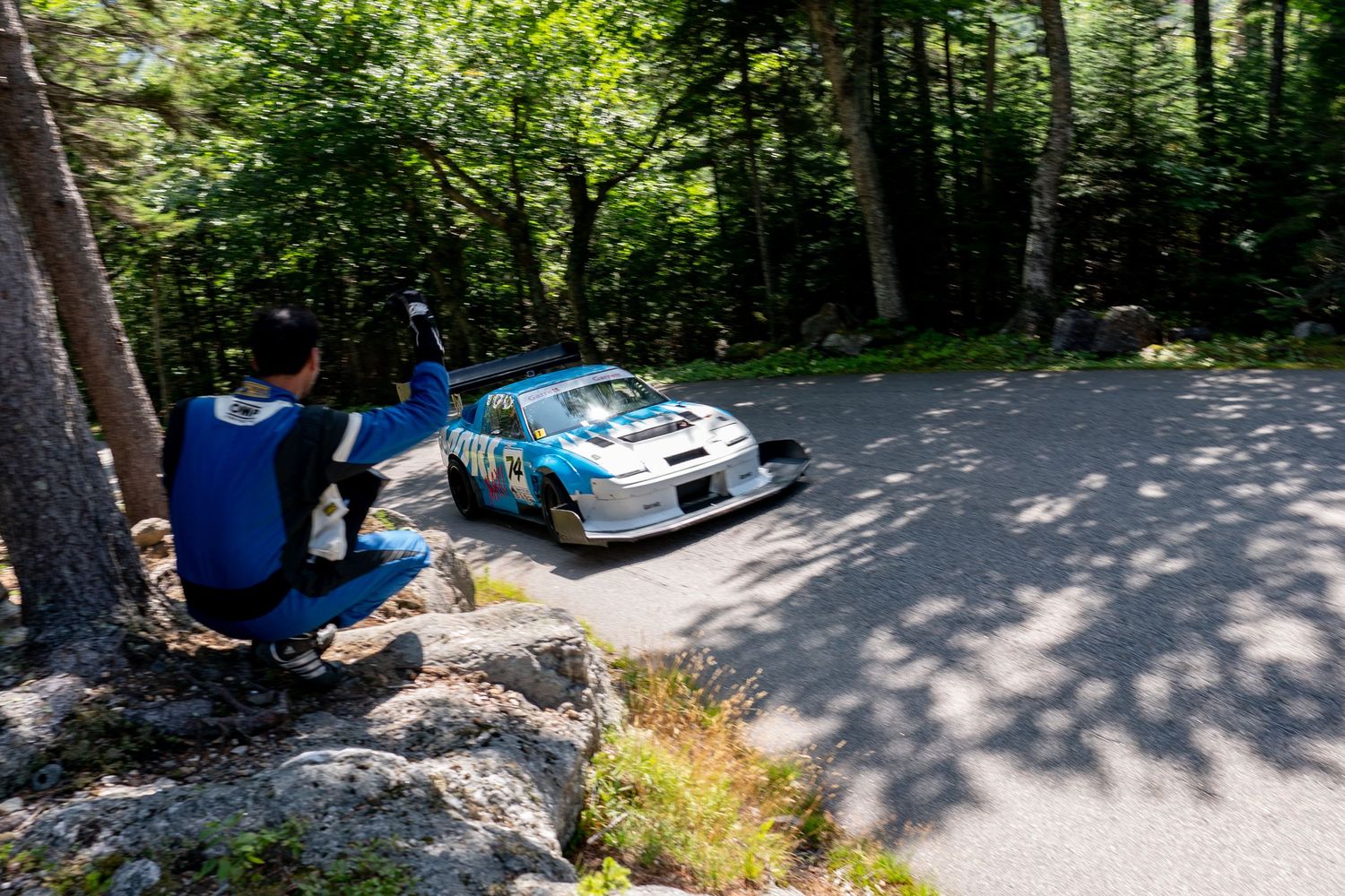 A rally car speeds past spectators on a mountain road during a race event.