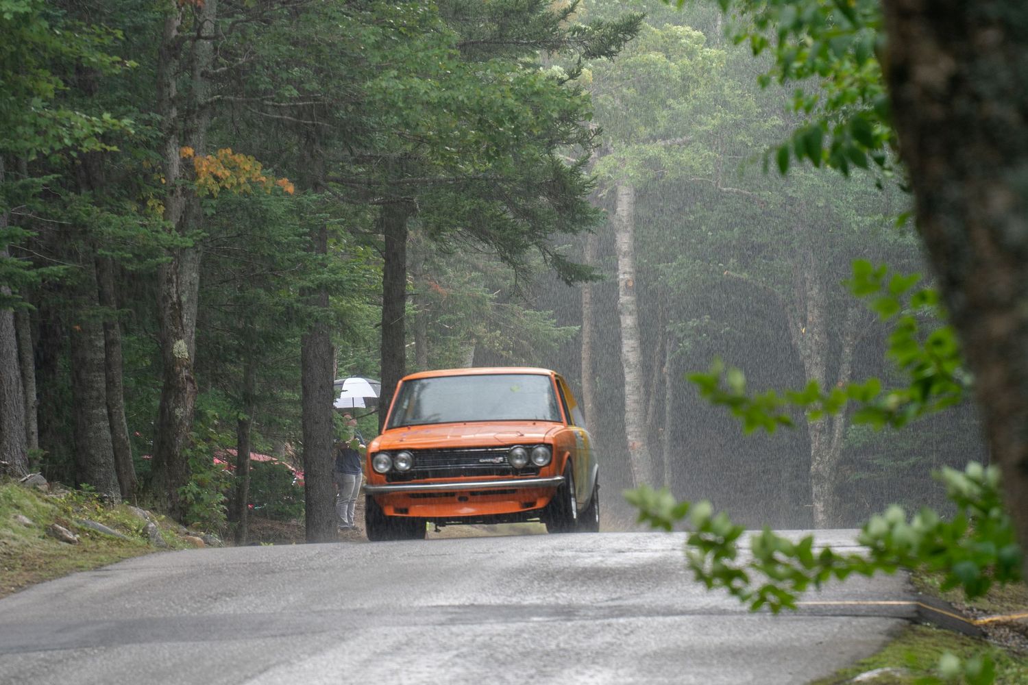 An orange vintage car catches air while cresting a hill on a misty forest road.