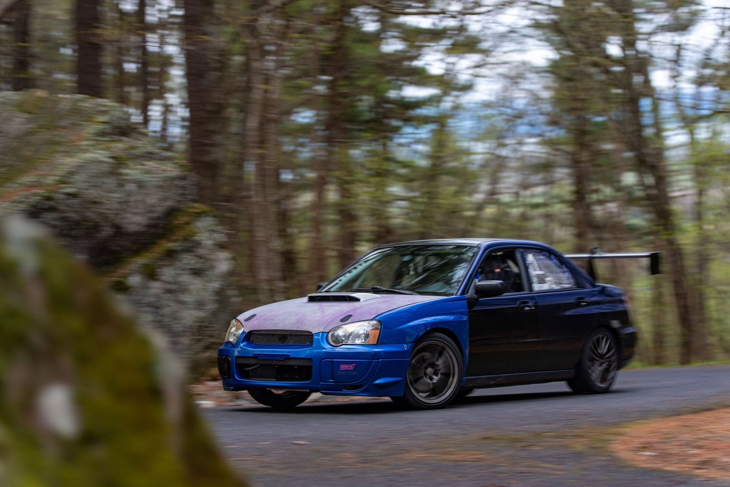 Blue Subaru WRX STI sedan with rear spoiler speeding on a winding forest road, motion blur background.