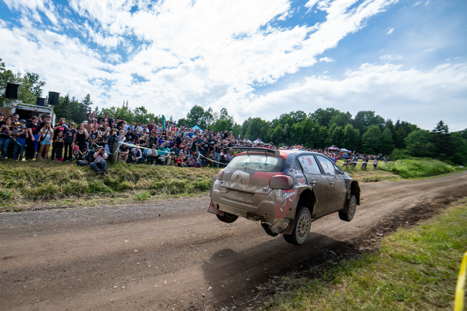Rally car jumping over a dirt road with spectators watching in the background.