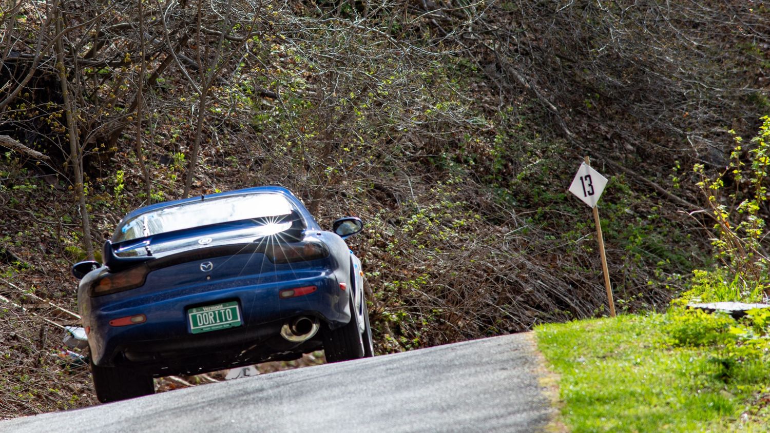 A dark blue sports car crests a steep hill on a narrow road surrounded by trees and a road sign.