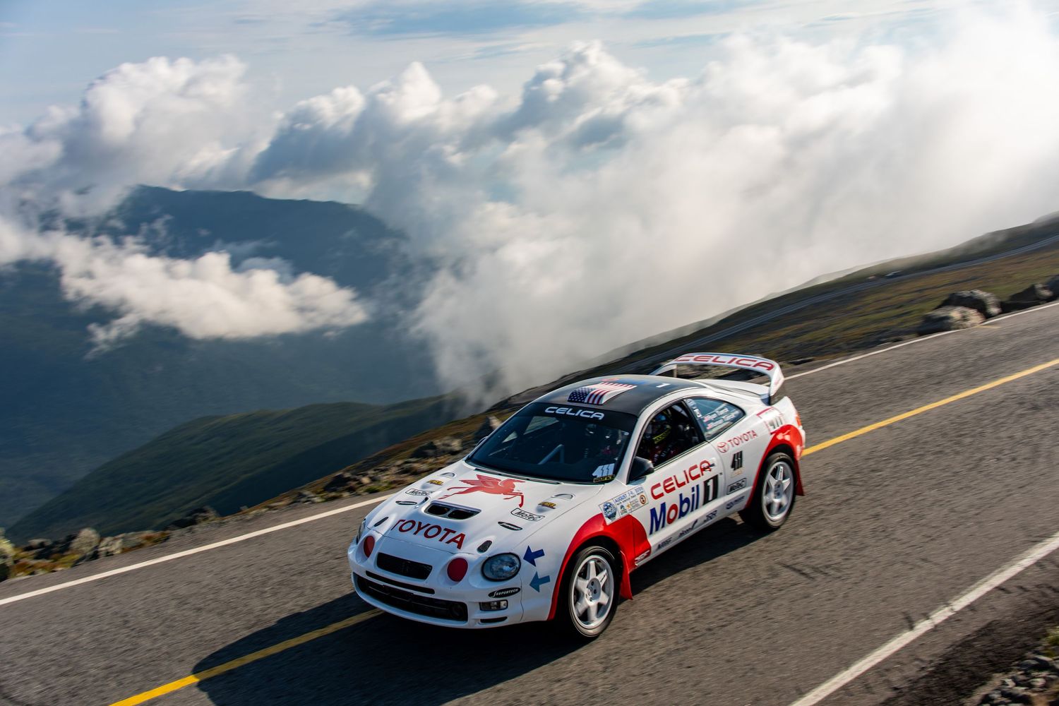 White Toyota Celica rally car racing on mountain road above clouds at Pikes Peak Hill Climb event.