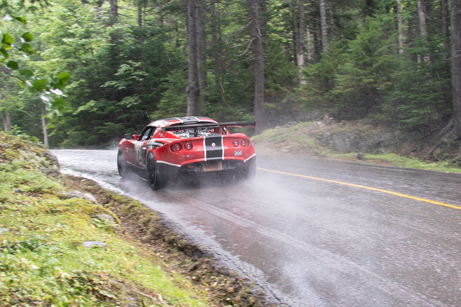 A red classic sports car slides through a wet corner on a rainy forest road.