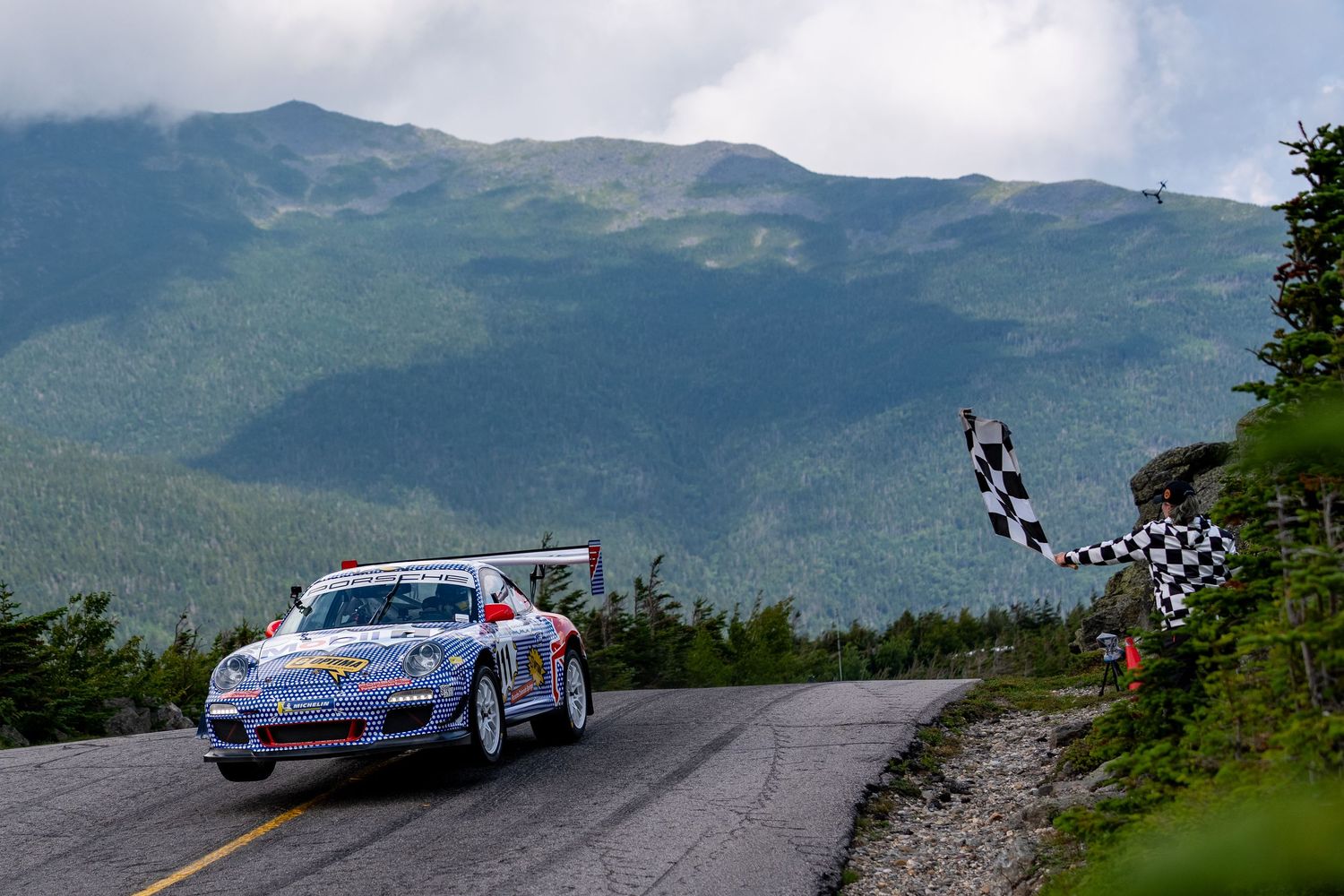 A Porsche race car crosses the finish line on a mountain road with a checkered flag and lush green peaks in the background.