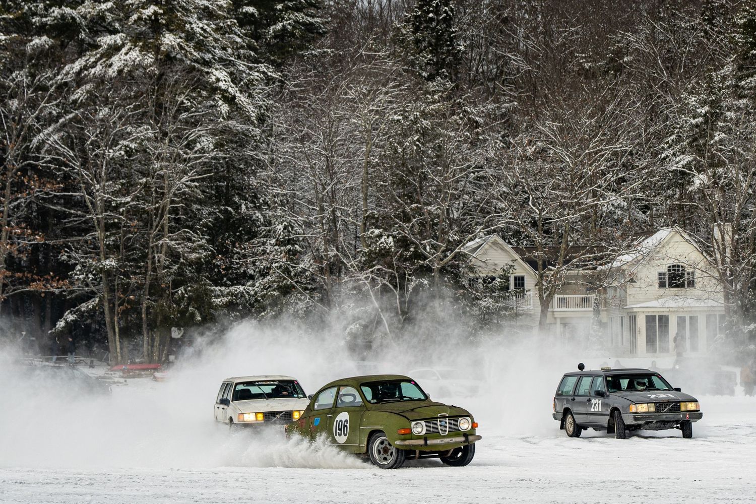 Classic cars racing through a snow-covered forest course during a winter competition.