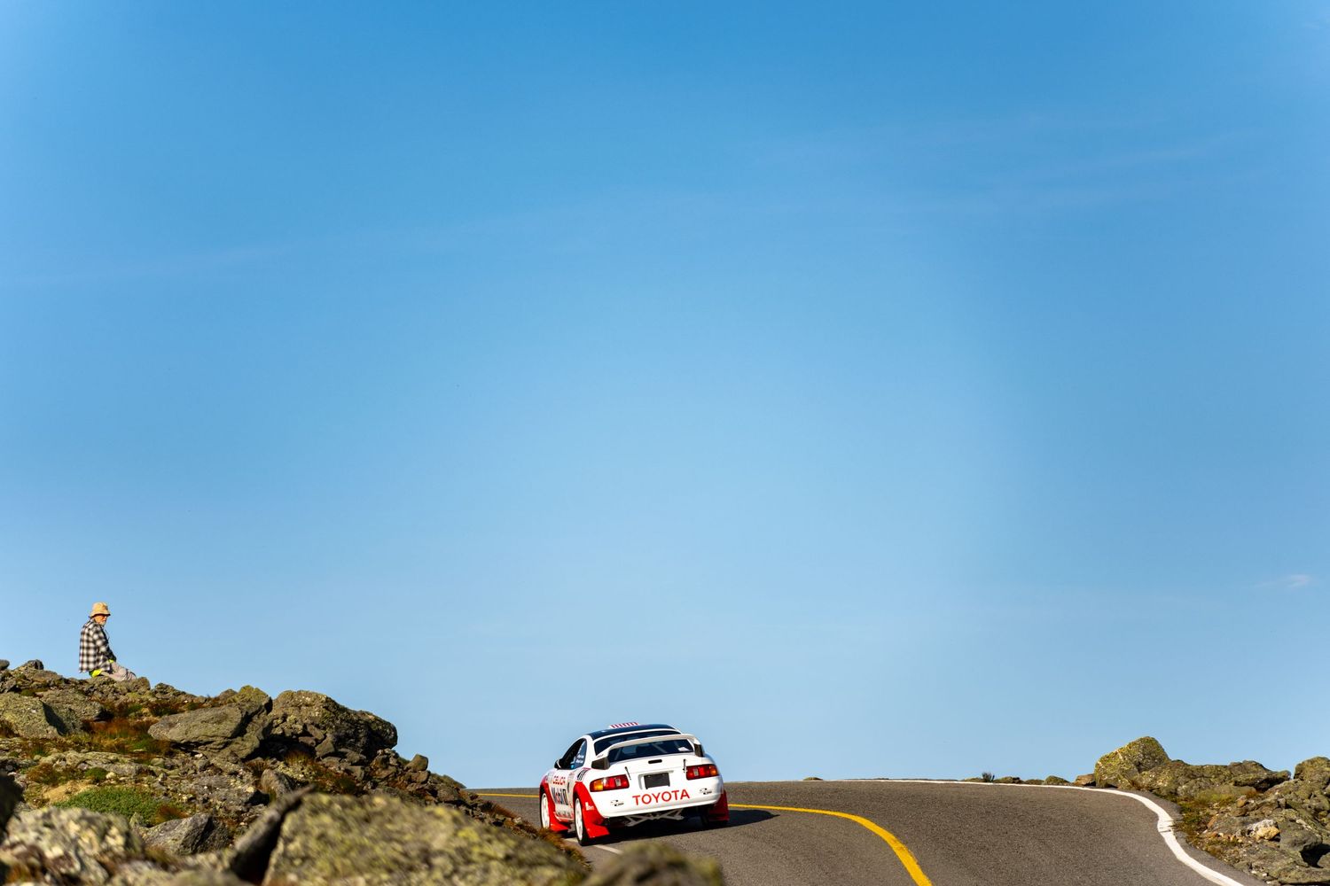 A red and white rally car drives along a winding mountain road under a clear blue sky.