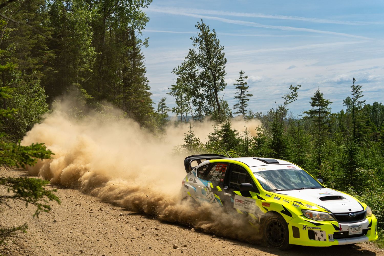 Rally cars race across dirt roads through scenic mountain forest terrain.