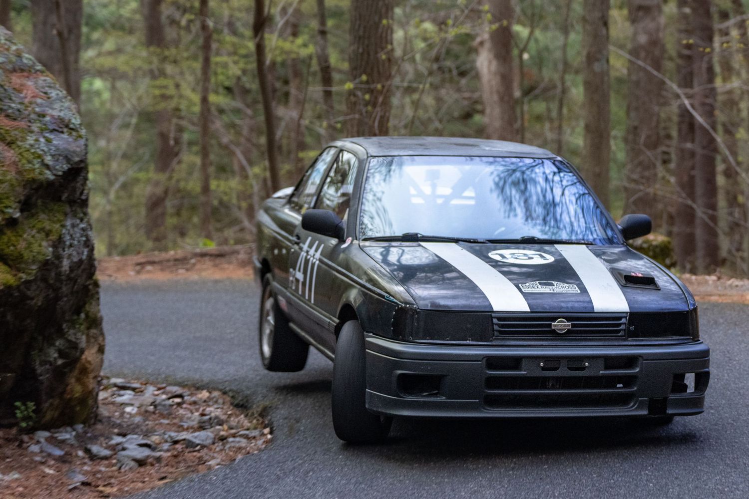 Black rally car with white stripes racing on a forest road during a motorsport event.