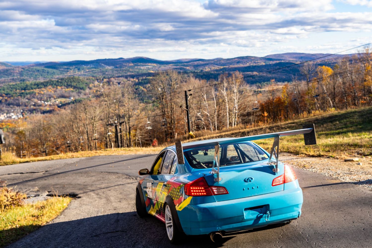A blue race car navigates a mountain road with scenic autumn valley views in the background.