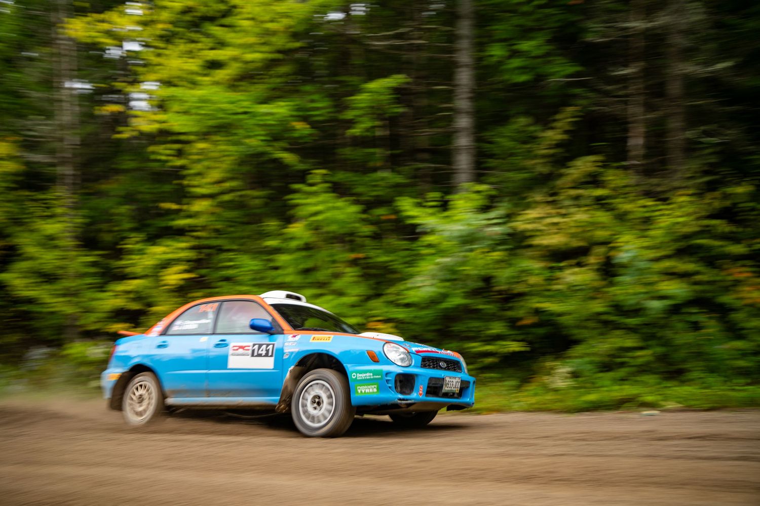 Blue rally car speeding through forest track during competitive race event.