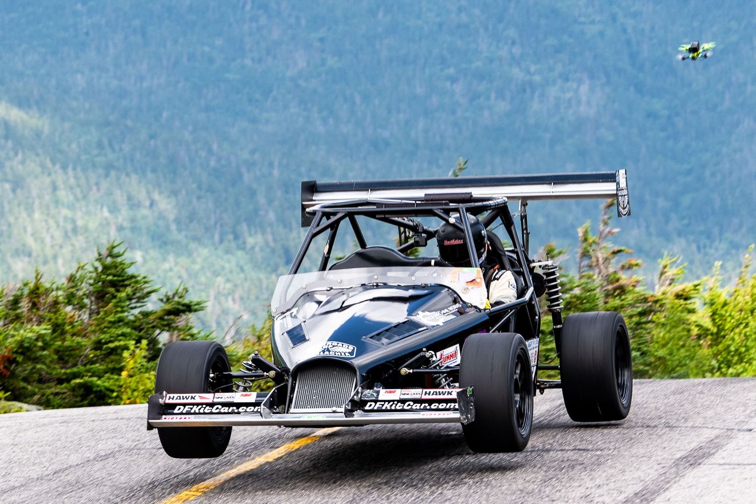 Open-wheel race car competing on a mountain hillclimb road course with forested peaks in the background.