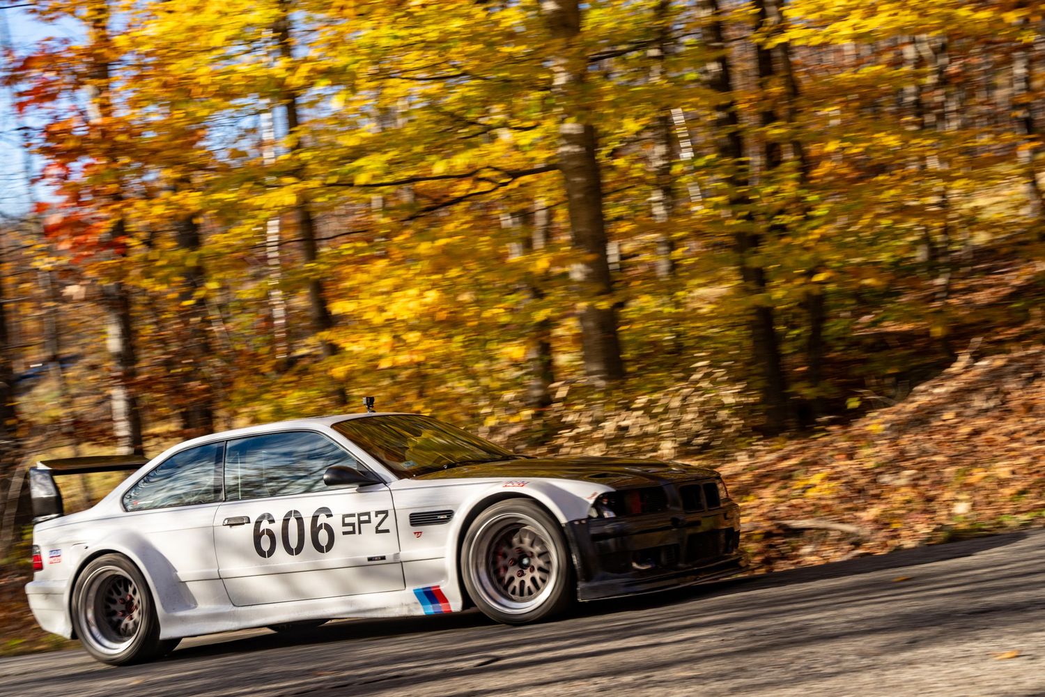 A white BMW race car drifts through fall foliage on a mountain course.