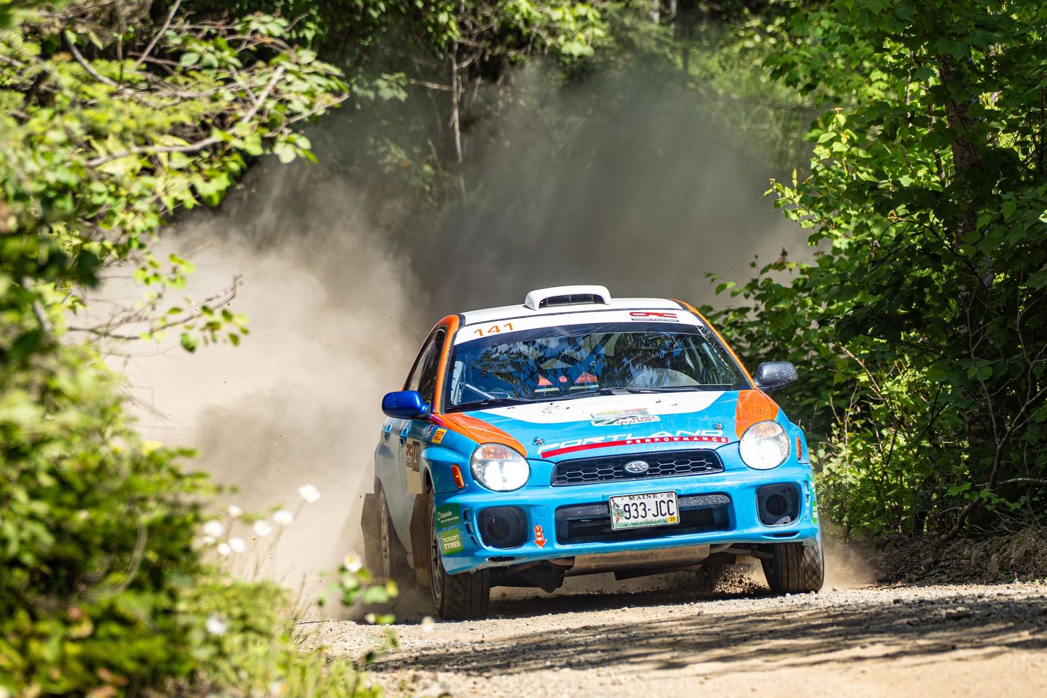 Rally cars drift through forest corners creating dramatic dust trails on a sunny day.