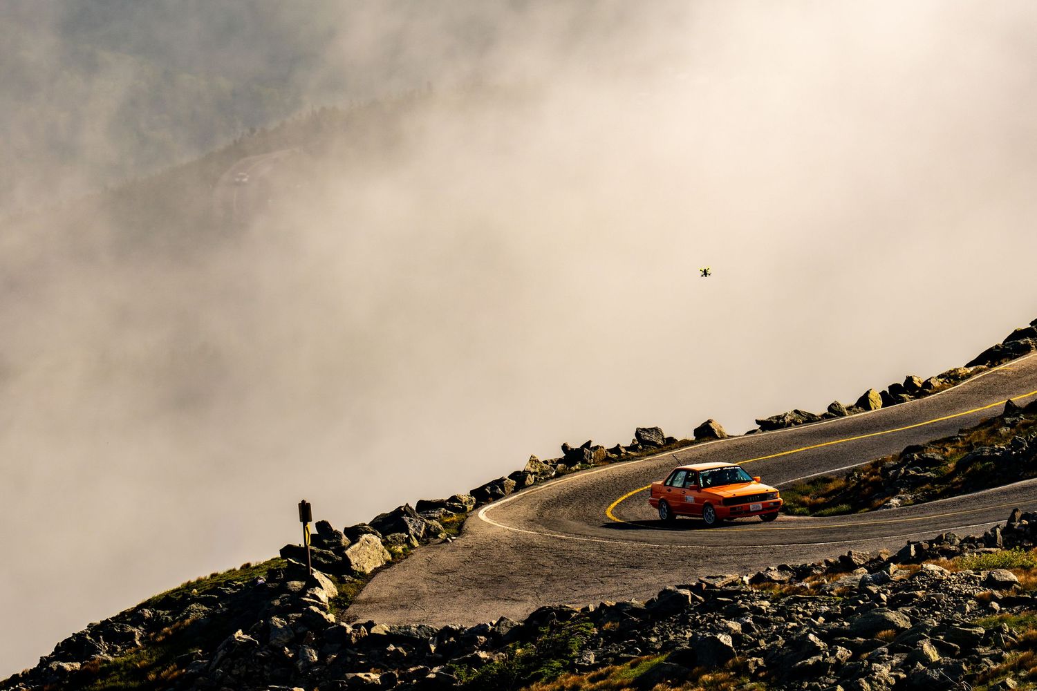 An orange sports car navigates a sharp mountain road curve surrounded by dramatic clouds and rocky terrain.