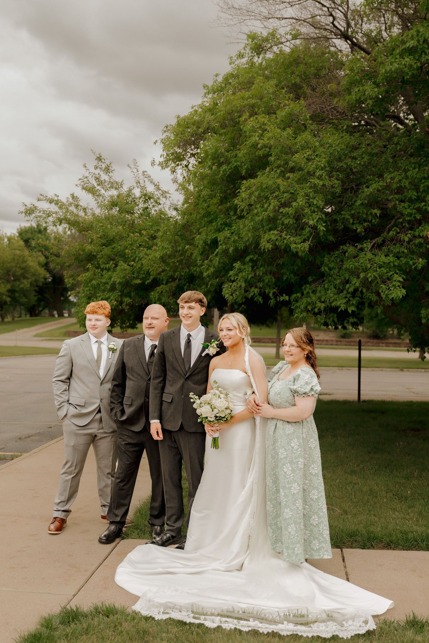 Wedding party group photo with bride in white gown and family members on sidewalk under cloudy sky.