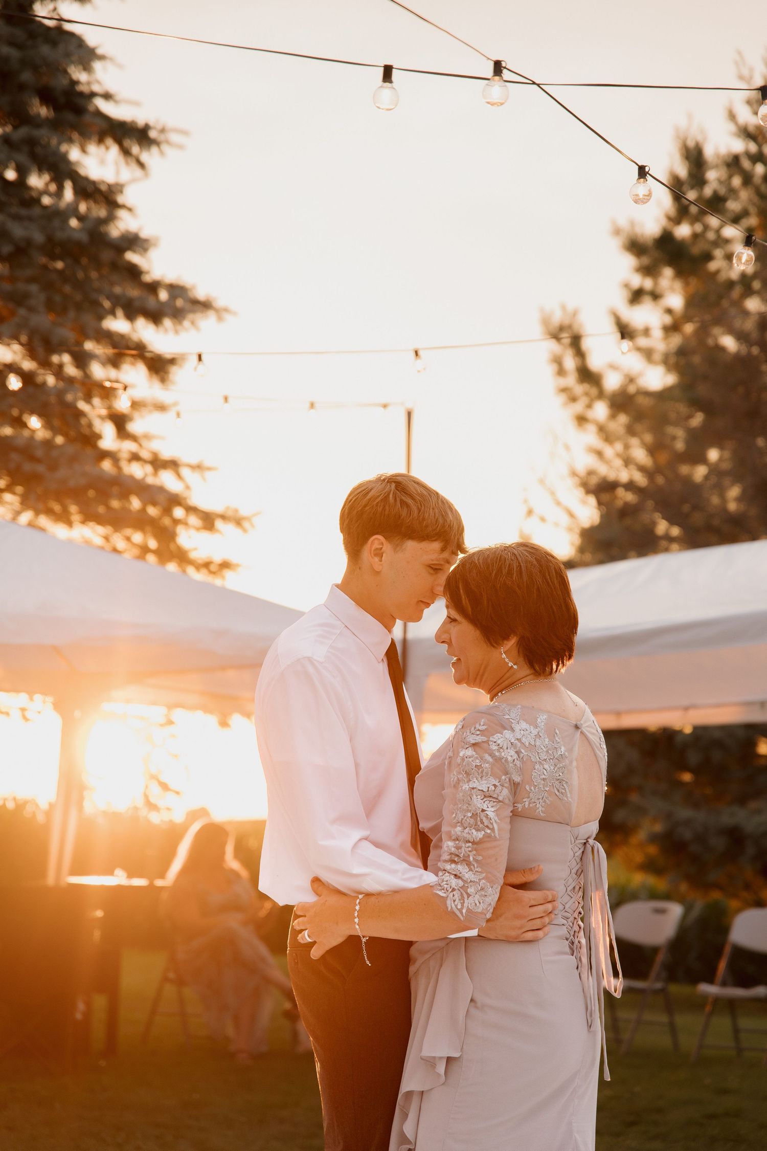 Couple sharing a romantic dance outdoors at golden hour wedding reception with string lights and white tent.