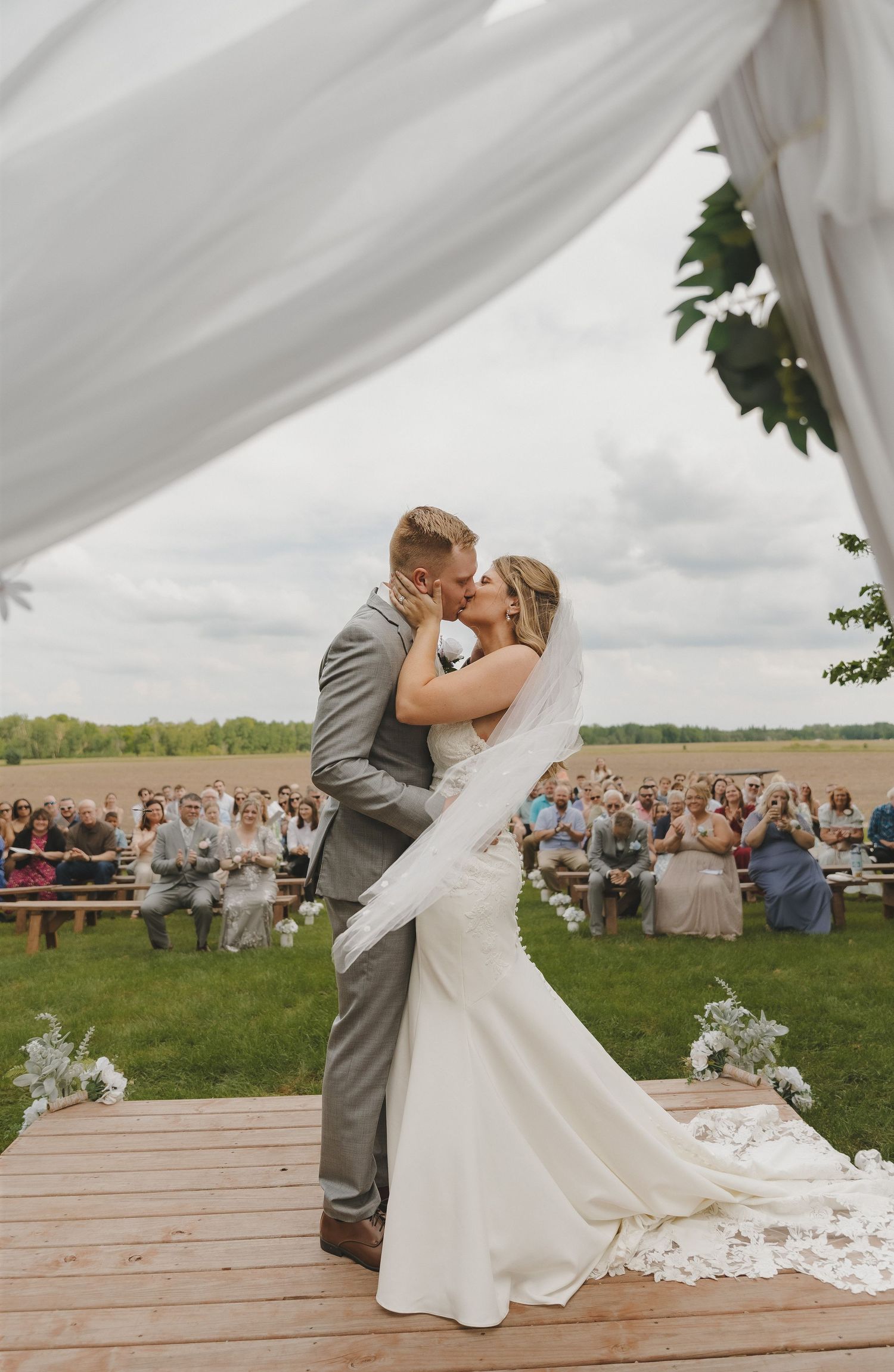 Bride and groom share first kiss on wooden altar with flowing white drapes and guests seated outdoors.